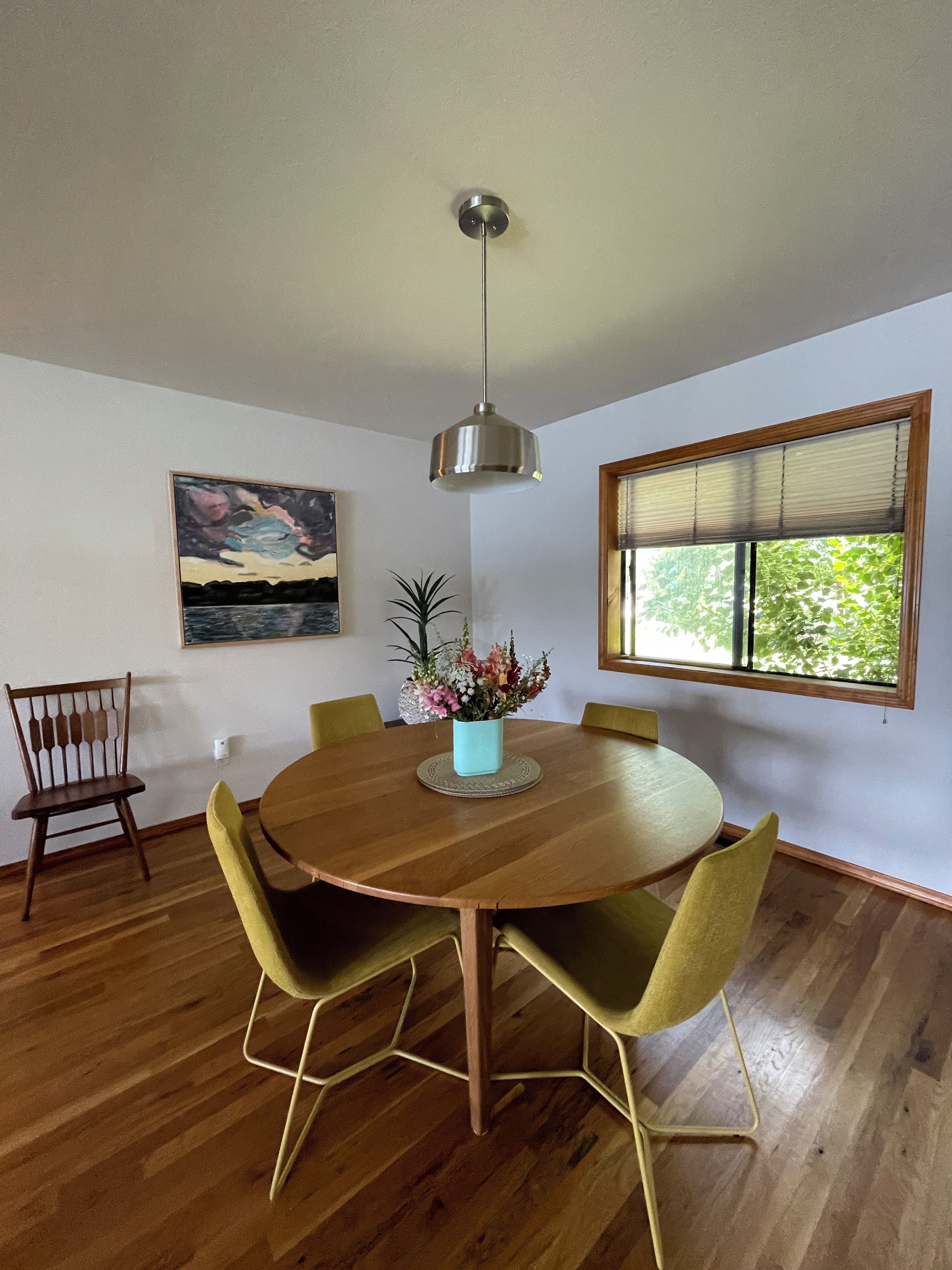 A dining area with a round wooden table, yellow chairs, a hanging light fixture, a window with blinds, and a painting on the wall.