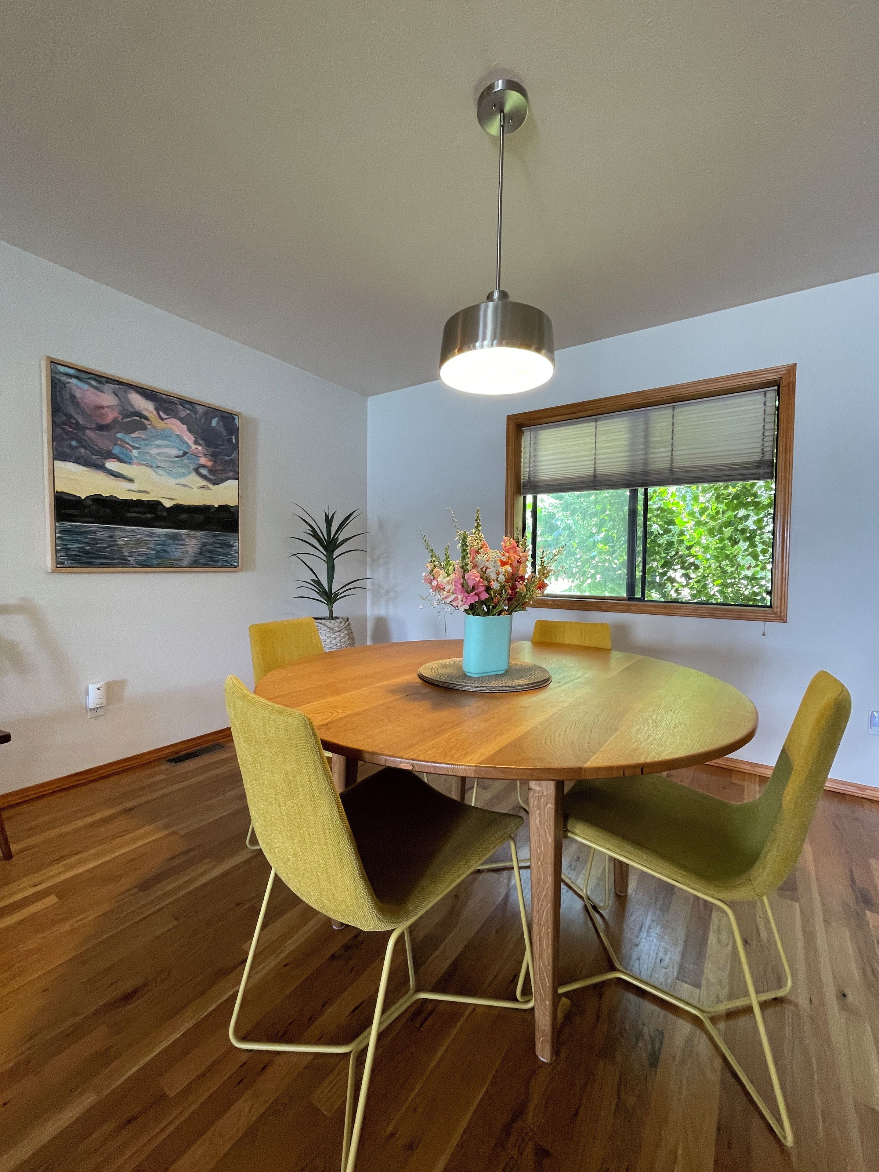 A dining area with a round wooden table, green upholstered chairs, a vase with flowers, and a painting on the wall.