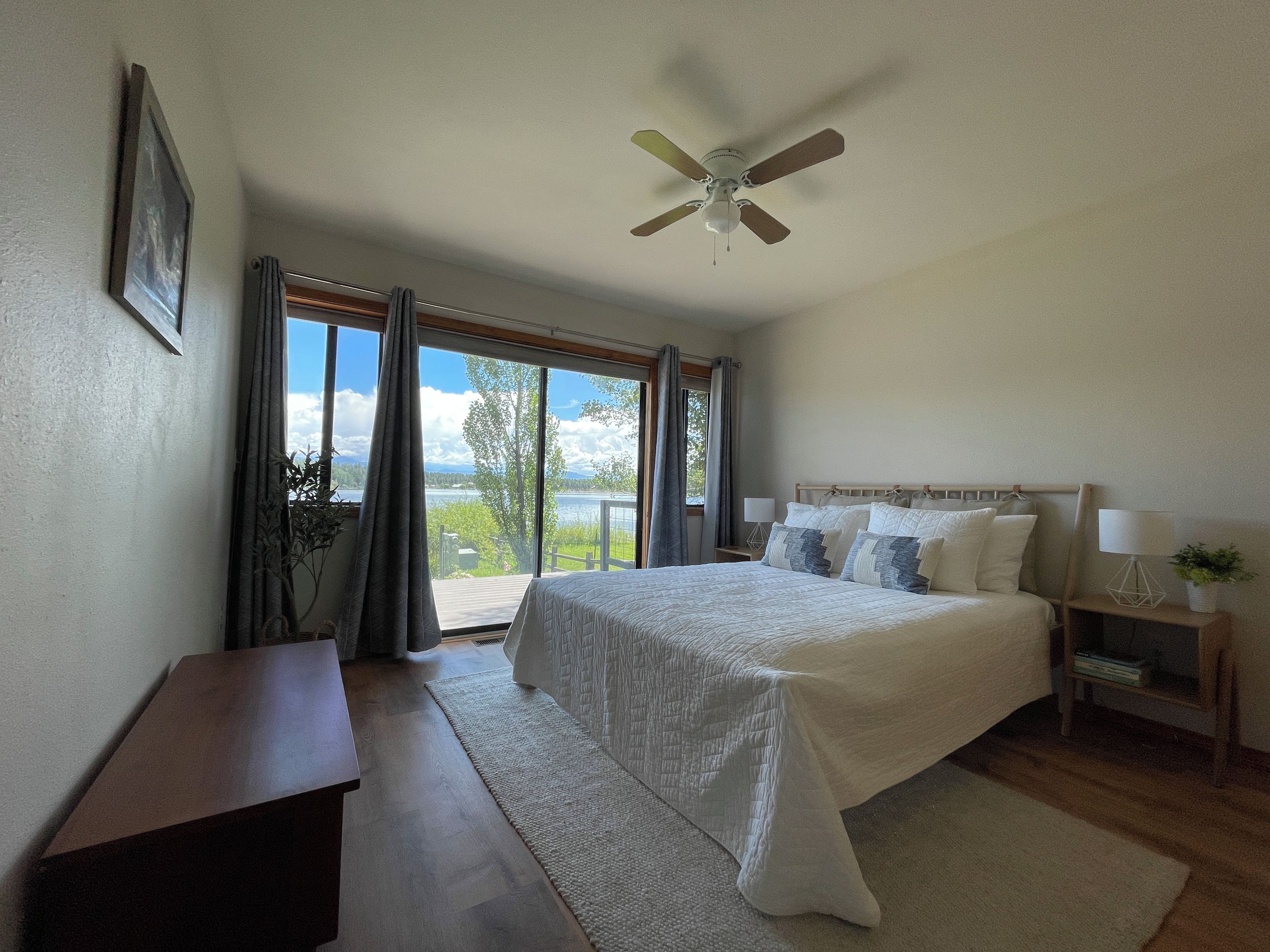 A bedroom with a bed, bedside tables, a ceiling fan, and a view of a lake through large windows.