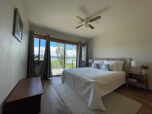 Un dormitorio con cama, mesitas de noche, ventilador de techo y vista a un lago a través de amplias ventanas.