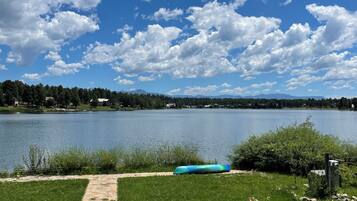 A lakeside patio with stone flooring, two Adirondack chairs, a canoe, and a grassy area with flowers.
