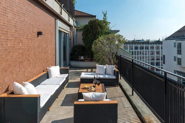 Una terraza en la azotea con mobiliario exterior, una mesa con una botella y vasos, y vistas de edificios circundantes.