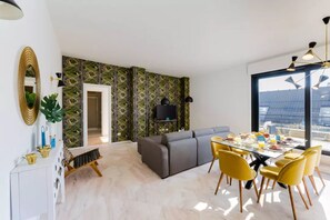 Una sala de estar moderna con un sofá gris, sillas amarillas y una mesa de comedor lista para el desayuno. Un ventanal ofrece vistas del paisaje urbano.