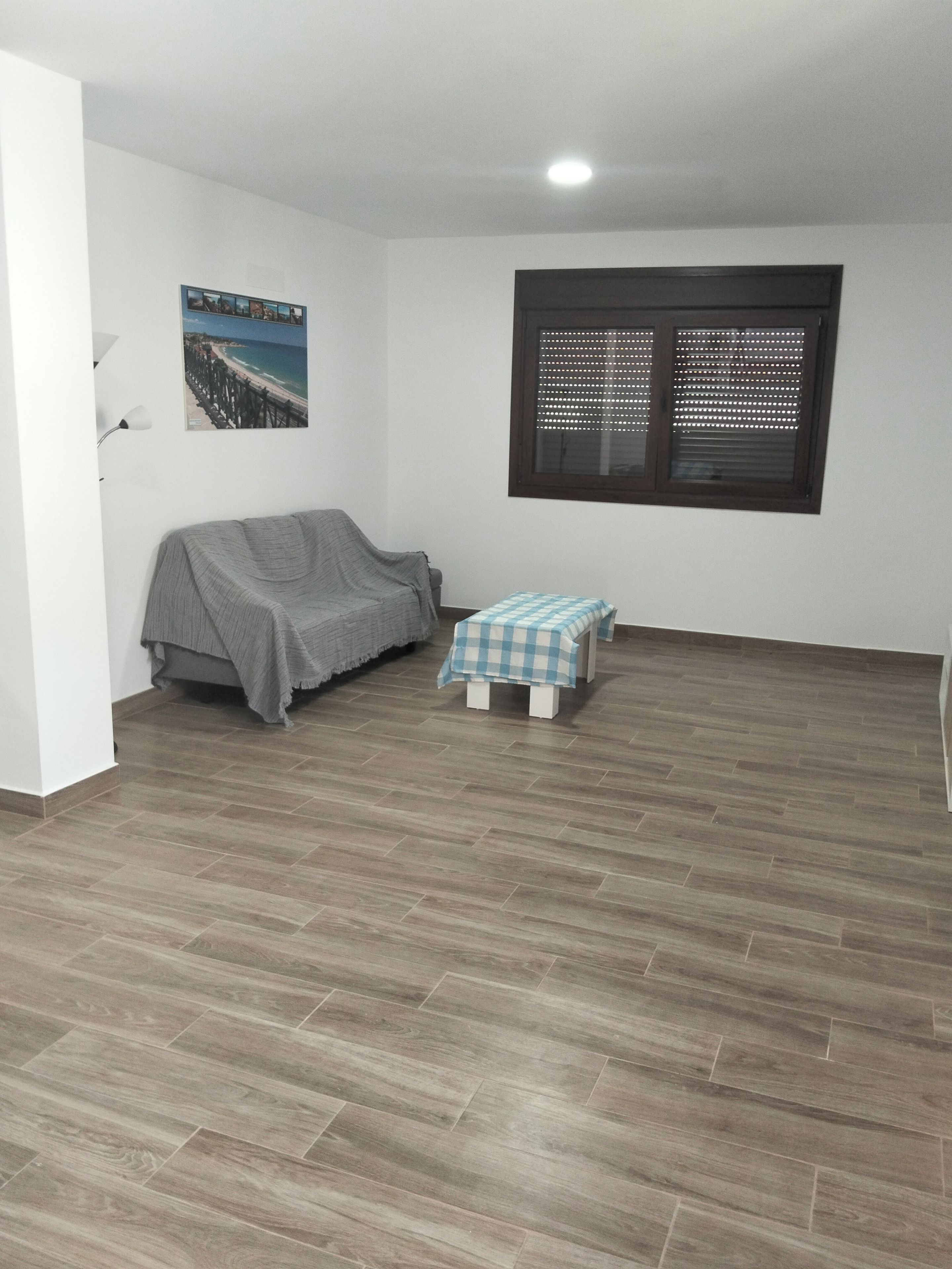 Une pièce avec un lit, une petite table, une fenêtre avec des stores et un tableau au mur.