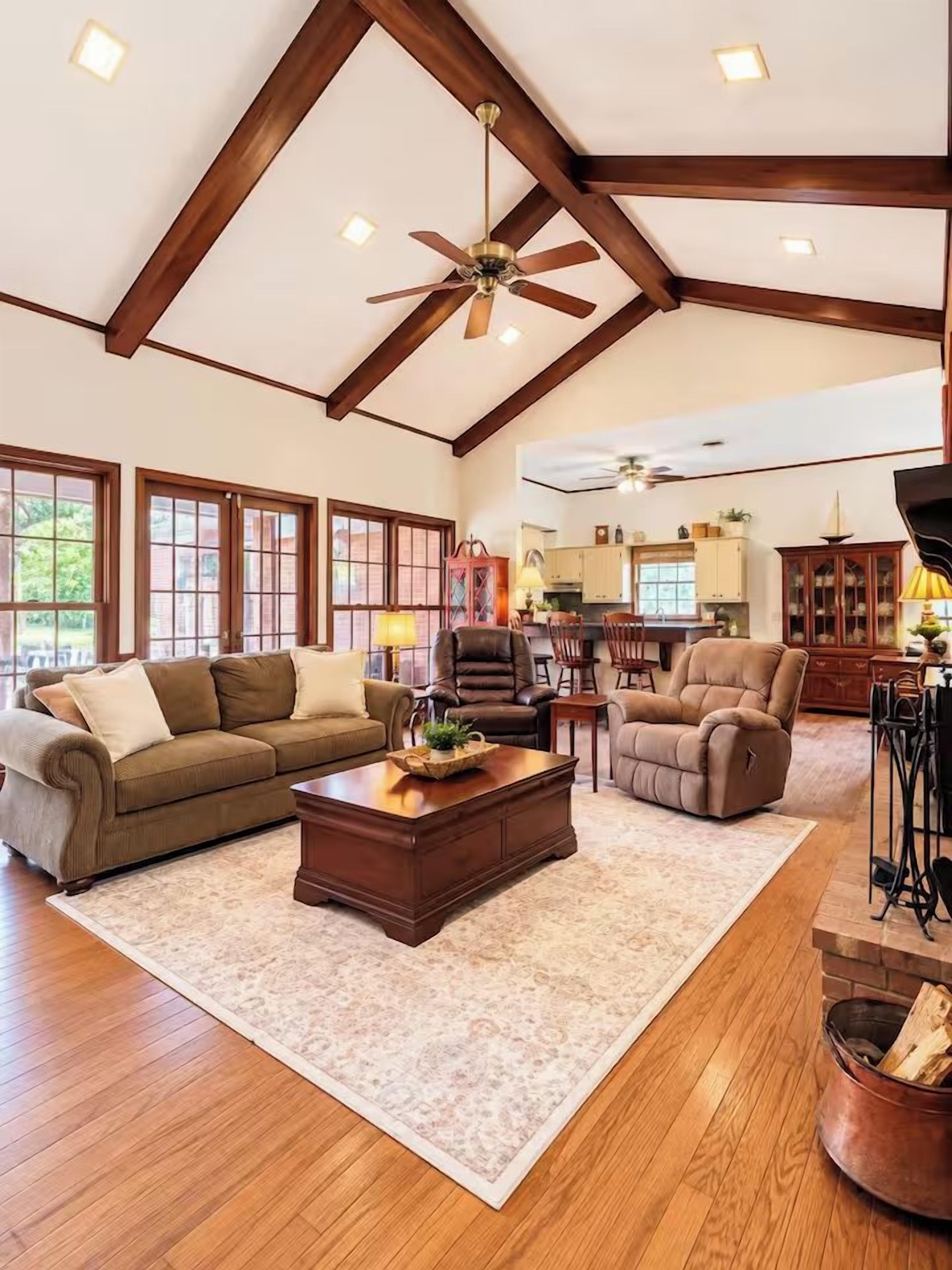 A spacious living room with a high ceiling, wooden beams, a ceiling fan, and a fireplace.