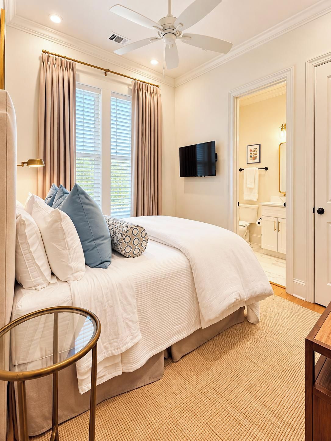 A bedroom with a bed, curtains, a ceiling fan, a TV, and a bathroom visible through an open door.