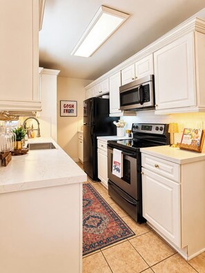 A kitchen with white cabinets, a black refrigerator, a microwave, and an oven. There is a rug on the floor and a framed picture on the wall.