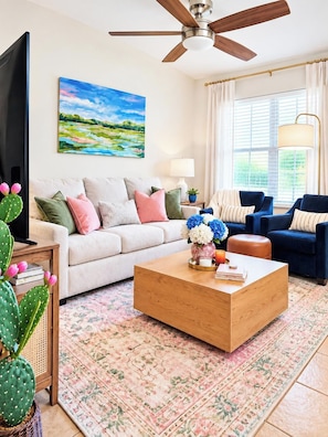 A living room with a sofa, two armchairs, a coffee table, a TV, a ceiling fan, a painting, a lamp, and a window with curtains.