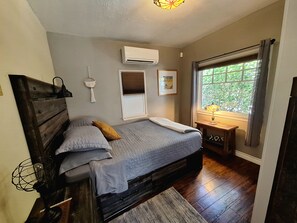 A bedroom with a bed, a bedside table, an air conditioning unit, a window with curtains, and a wall-mounted lamp. - Charming 2-Bdrm Historic Bungalow~ Hummingbird Garden & HotTub in DT Fullerton! (Fullerton)