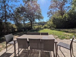 En udendørs terrasse med et bord og stole, omgivet af træer og græs.