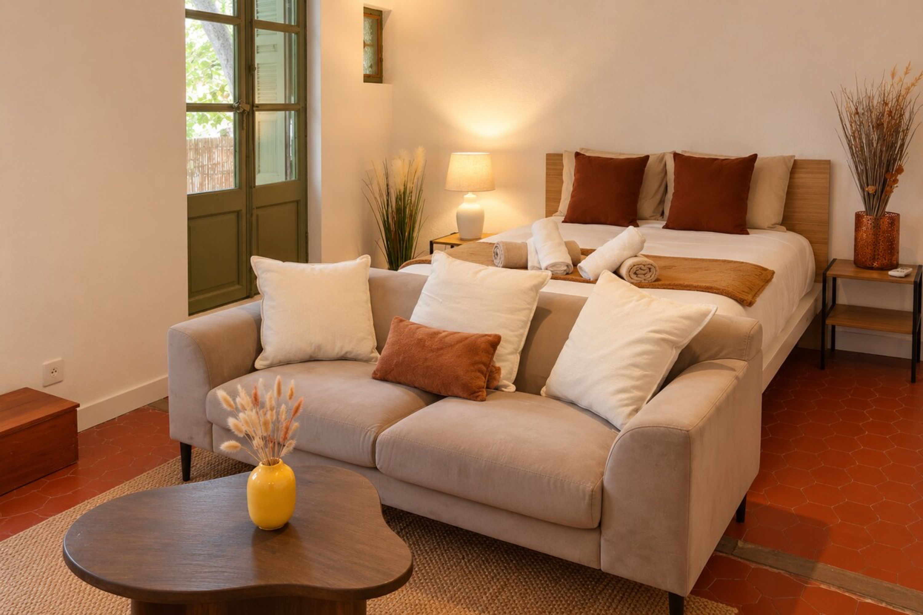 Une chambre d’hôtel avec un canapé, un lit avec plusieurs oreillers, une table de chevet avec une lampe et un vase contenant des plantes séchées.