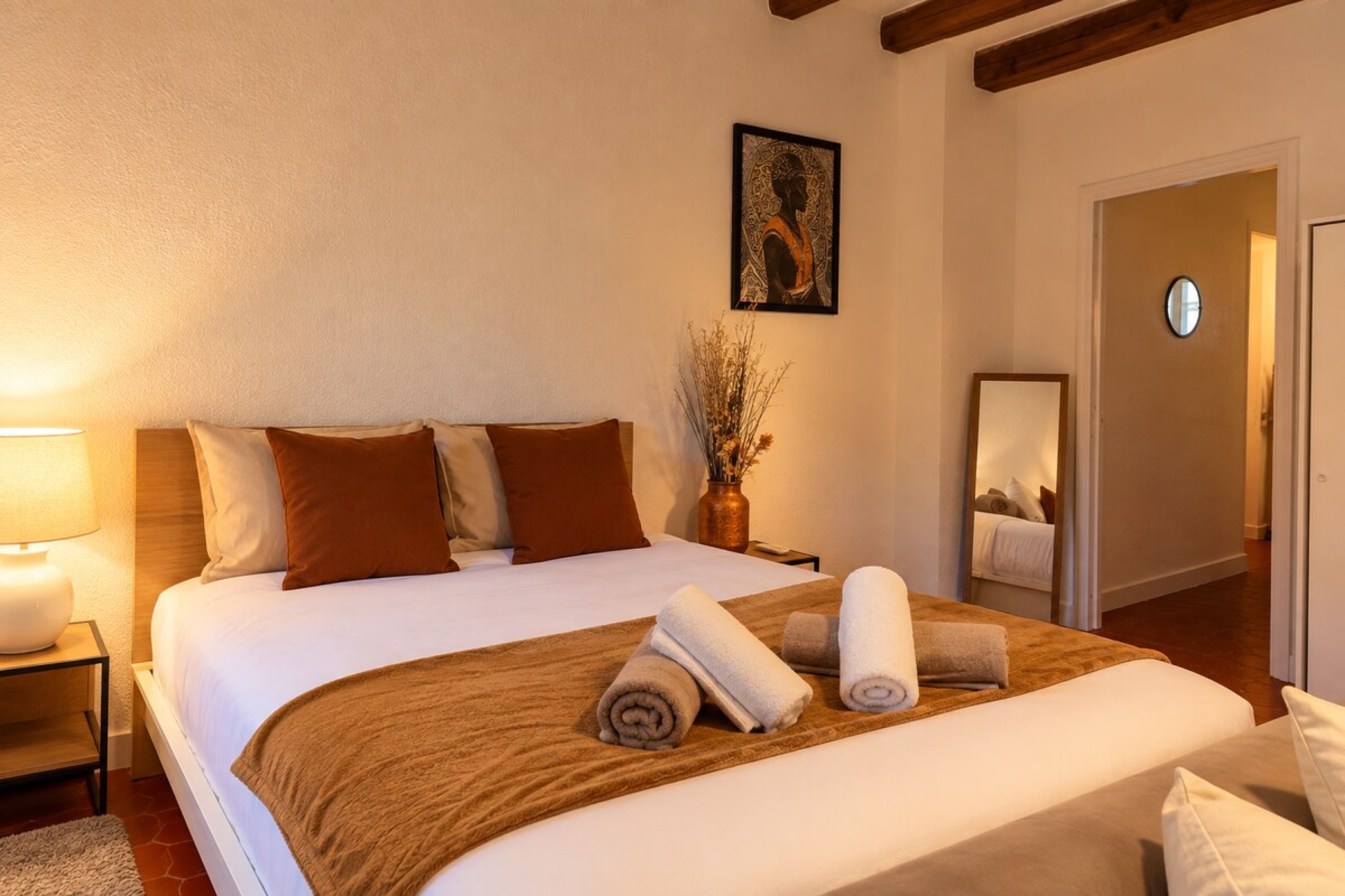 Una habitación de hotel con una cama grande, una mesita de noche, un florero con plantas secas y un cuadro en la pared.