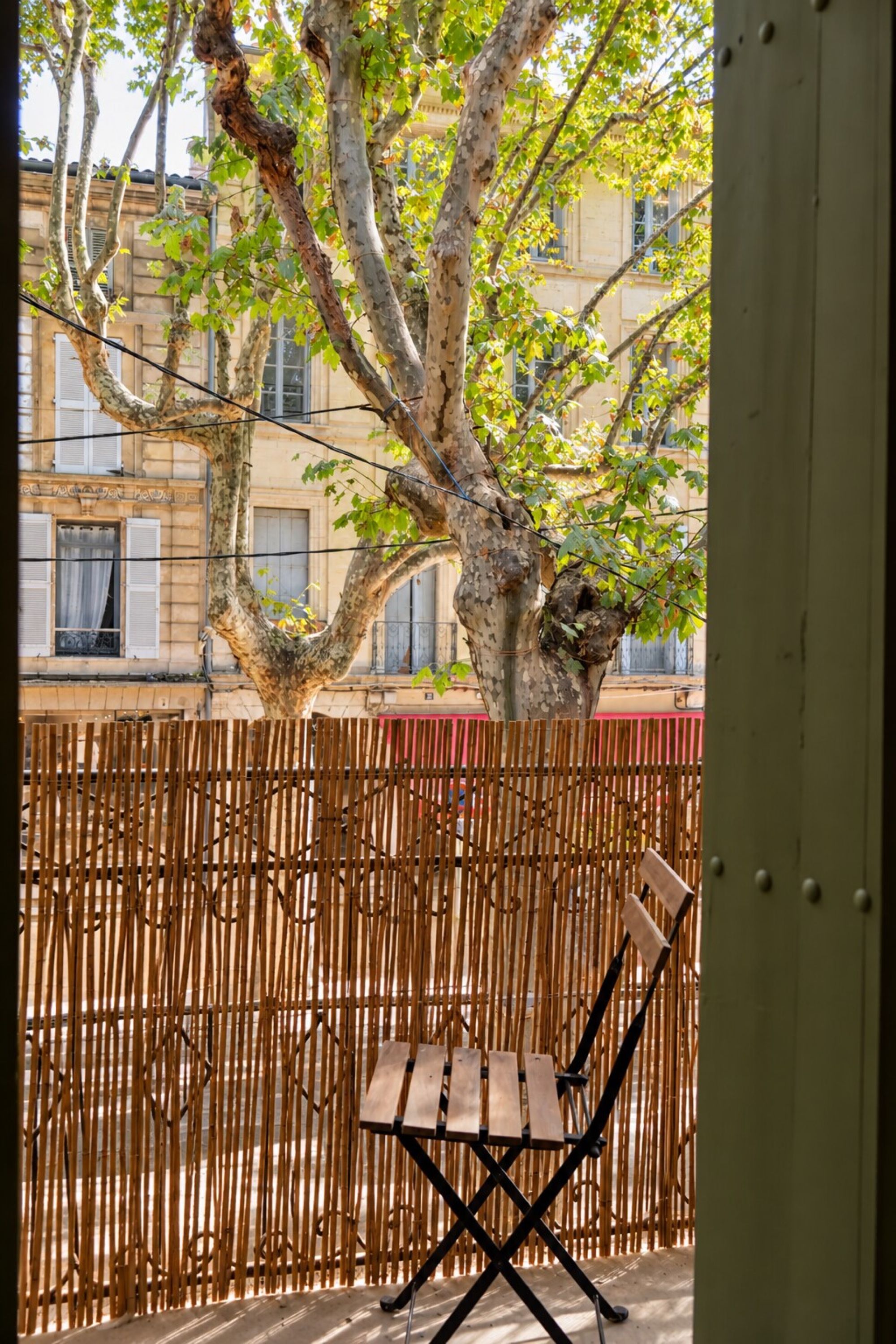 Una silla de madera frente a una valla de bambú, con un árbol y edificios al fondo.