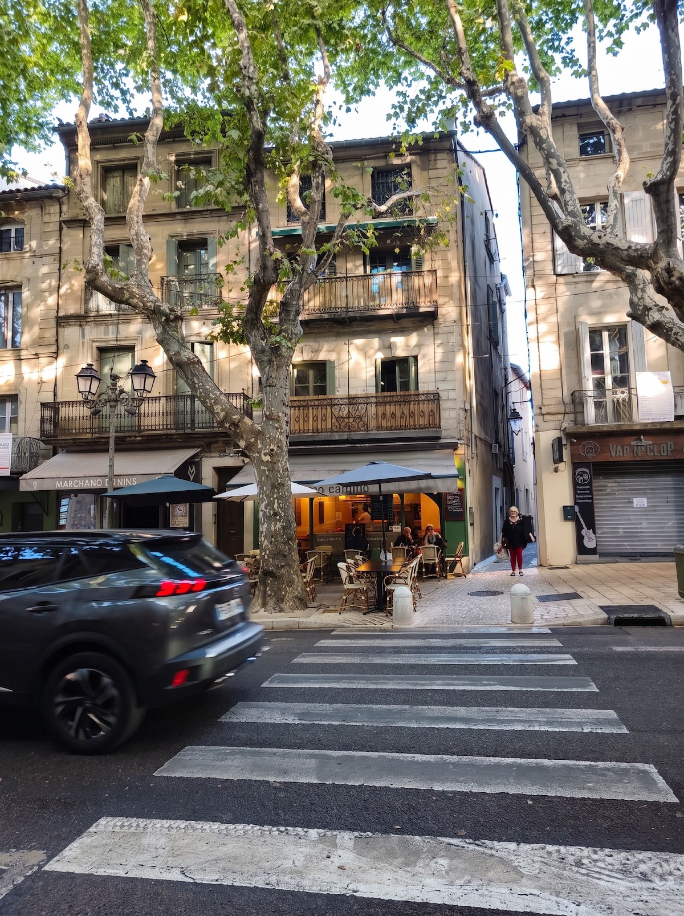 Une vue de rue avec un passage piéton, des arbres et un immeuble doté de balcons et d’une terrasse.