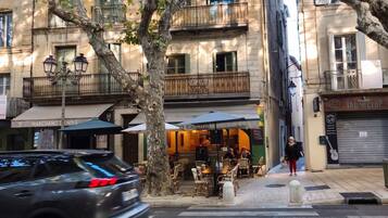 Vista de la calle con un paso de cebra, árboles y un edificio con balcones y terraza.
