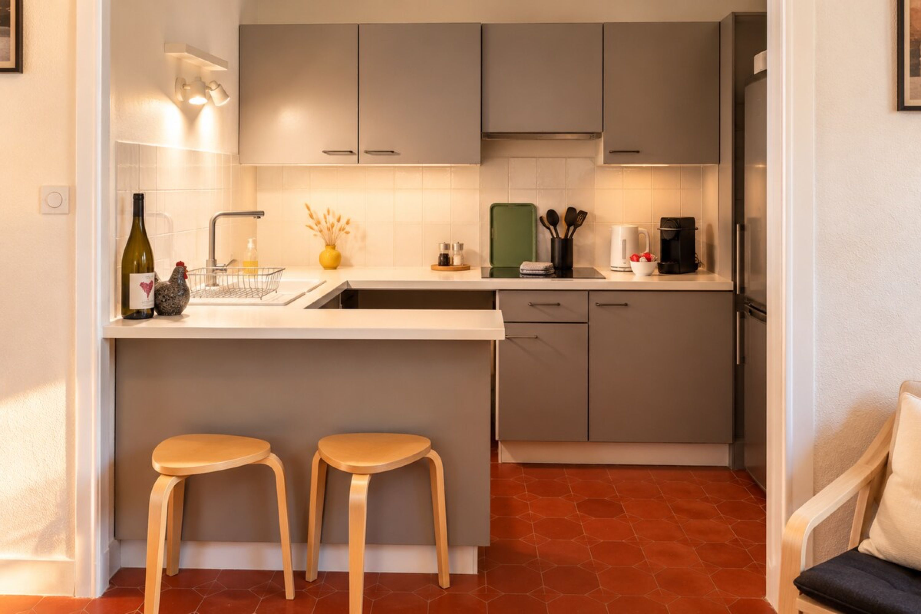 Une cuisine moderne avec des meubles gris, un plan de travail blanc, deux tabourets en bois et un lave-vaisselle encastré.