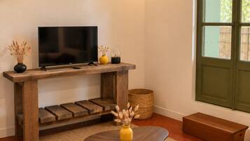 Una sala de estar con una televisión sobre un mueble de madera, un sofá, una mesa de centro y una ventana con vistas a zonas verdes.