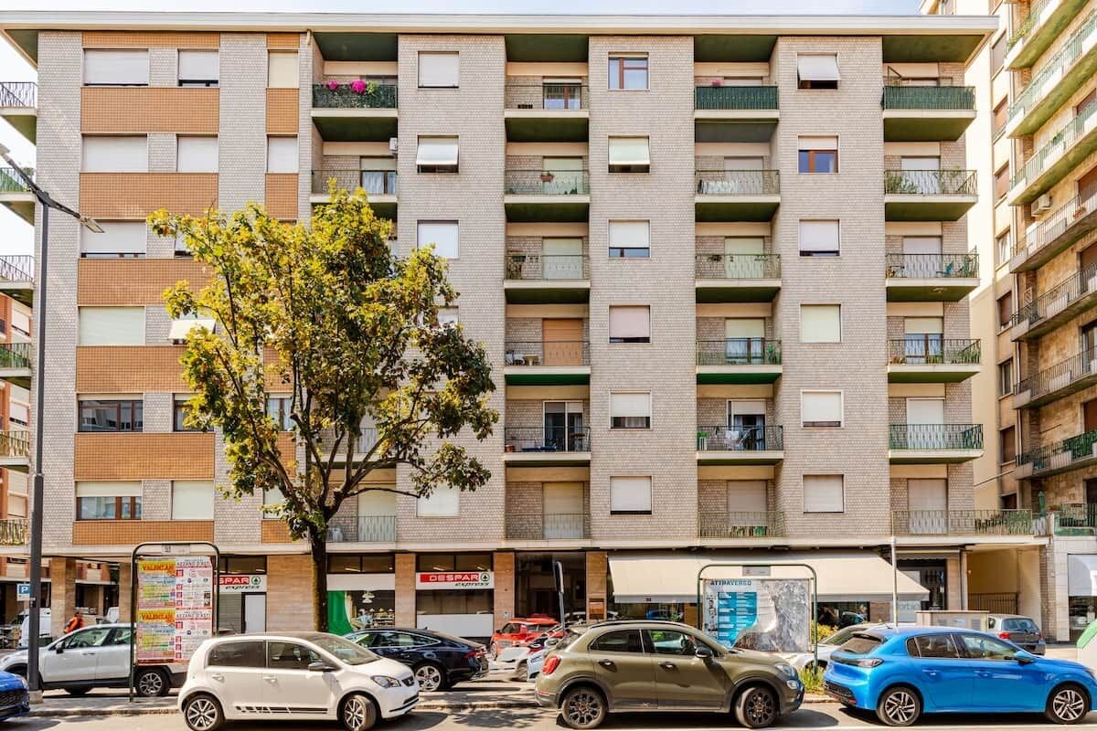 A multi-story residential building with balconies, a tree in front, and parked cars.