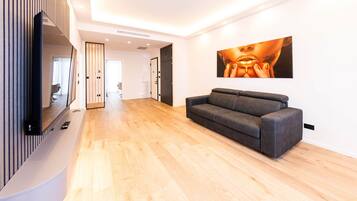 A modern living room with a grey sofa, wooden flooring, a flat-screen TV mounted on a wall, and a large framed artwork.