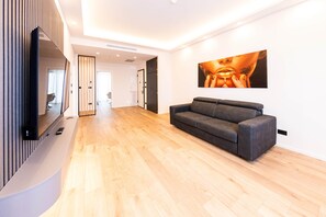 A modern living room with a grey sofa, wooden flooring, a flat-screen TV mounted on a wall, and a large framed artwork.