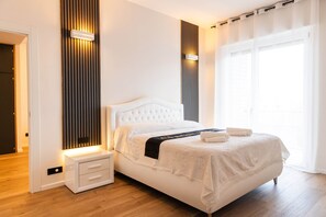 A modern bedroom with a white bed, a nightstand, a wall-mounted light, and a large window with curtains. - Charme Vittoria | Elegant Apartment in Downtown Asti (Asti)