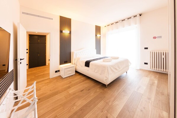 A modern bedroom with a bed, bedside tables, a nightstand, a radiator, and a door leading to another room.