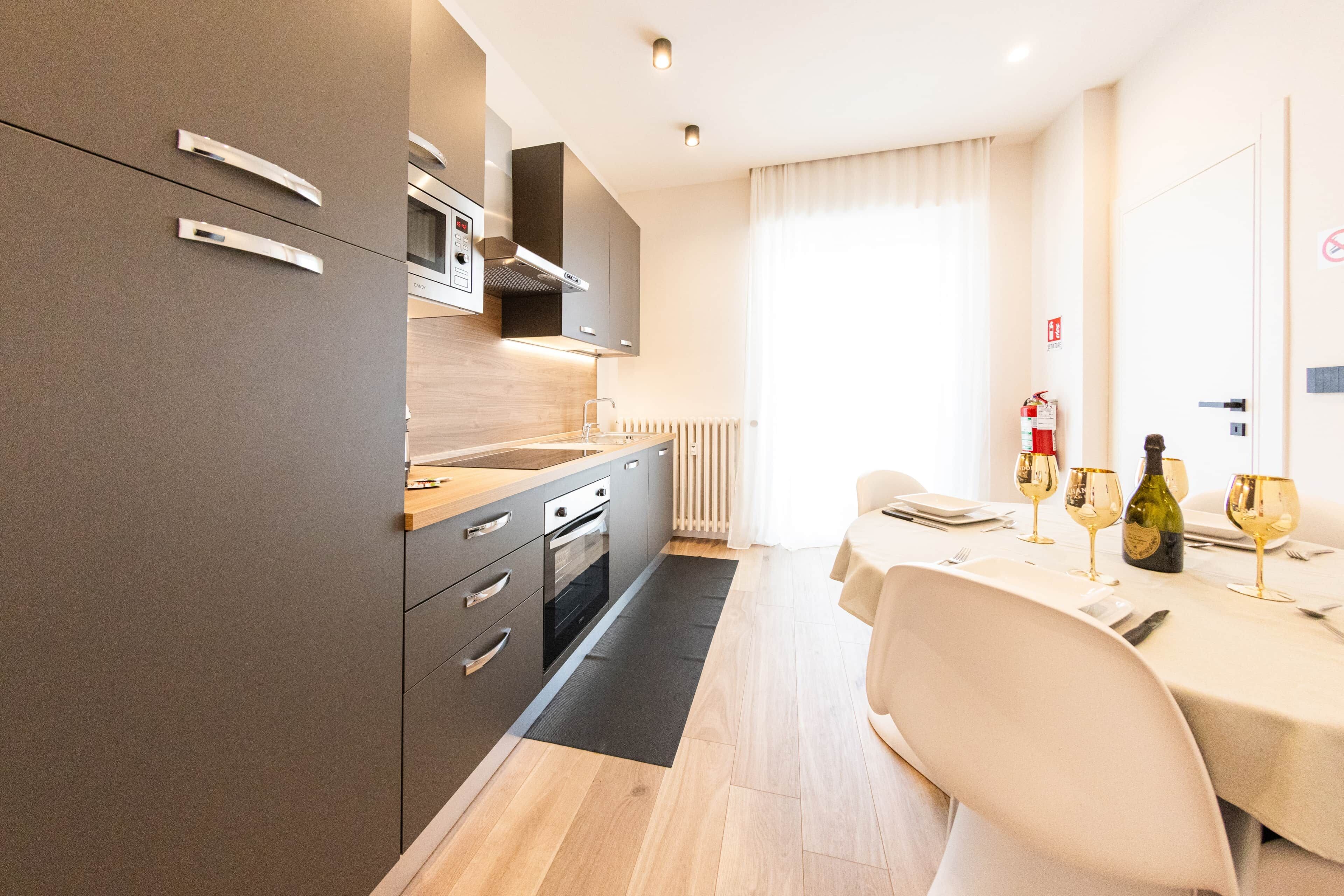 Een moderne keuken met een ingebouwde oven, een magnetron en een kookplaat. Er is een eethoek met een tafel gedekt voor twee personen, en een brandblusser bevestigd aan de muur.