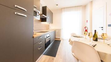 Een moderne keuken met een ingebouwde oven, een magnetron en een kookplaat. Er is een eethoek met een tafel gedekt voor twee personen, en een brandblusser bevestigd aan de muur.