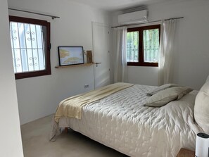 Ein Schlafzimmer mit einem Bett, einem Fenster mit Vorhängen, einer Wand-Klimaanlage und einem Regal mit einem gerahmten Bild.