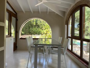 A dining area with a glass table and white chairs, large arched windows with a view of greenery, and a ceiling fan. - New traditional-style villa just a stone's throw from a beautiful cove (Jávea)