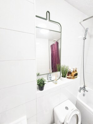 A modern bathroom with a white tiled wall, a glass shower enclosure, a white toilet, and a mirror with a hanging towel rack. - Elegant 2 Bedroom Apartment in Erith Breathtaking River Thames View (Erith)
