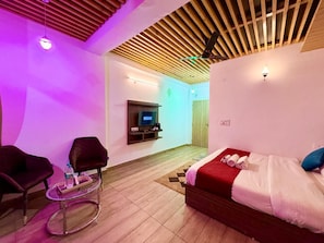 A hotel room with a bed, a TV mounted on the wall, a small table with a chair, and a wooden ceiling.