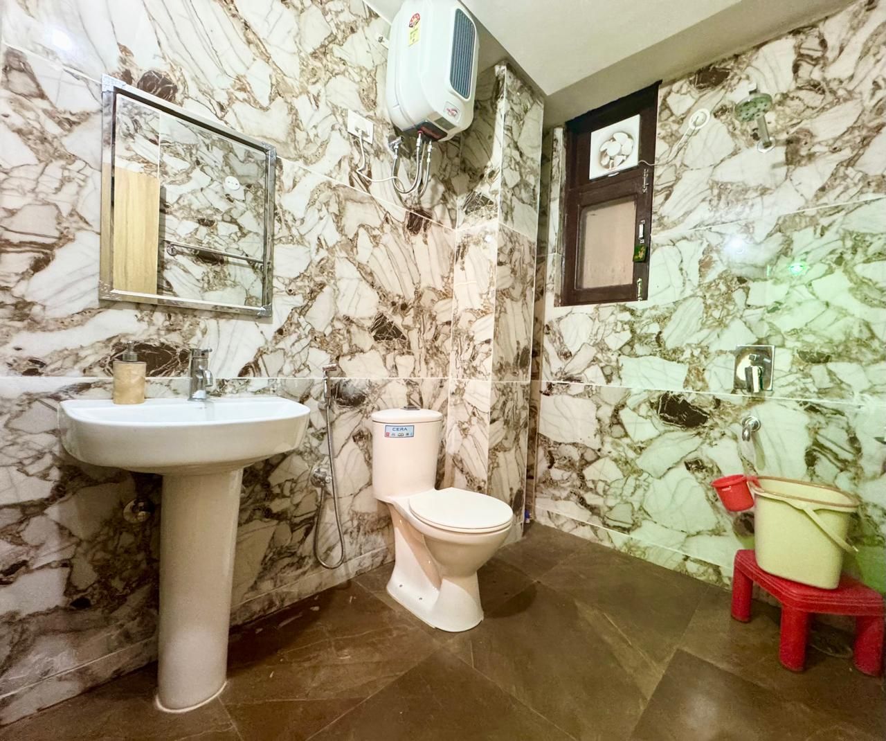 A bathroom with a marble wall, a white sink, a toilet, and a red stool.