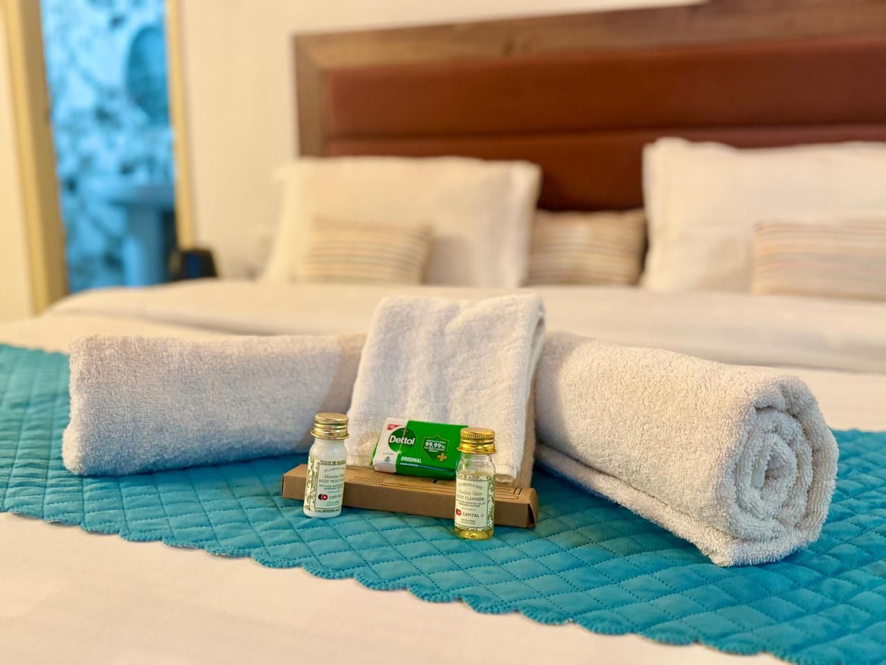 Una cama bien tendida con toallas blancas enrolladas y una pequeña bandeja con dos botellas y una caja verde.