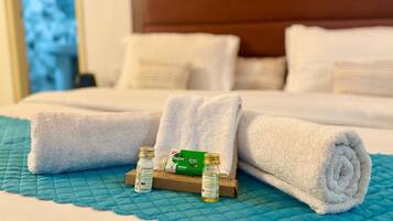Una cama bien tendida con toallas blancas enrolladas y una pequeña bandeja con dos botellas y una caja verde.