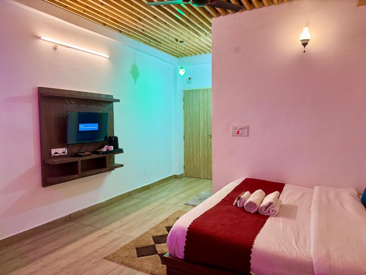 A hotel room with a bed, a flat-screen TV mounted on the wall, a wooden ceiling, and a door in the background.