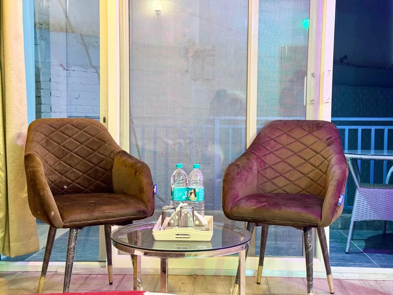 Two armchairs with a quilted pattern, a glass table with a tray of bottled water, and a wicker chair in the background.