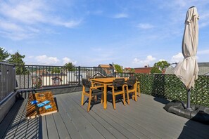 A rooftop deck with a wooden table and chairs, a cushioned seating area, and a pergola with an umbrella. - Modern 4BR Container Home | Epic Private Rooftop (Pittsburgh)