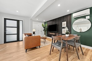 A modern living room with a sofa, dining table, chairs, a TV, and an air conditioning unit. - Modern 4BR Container Home | Epic Private Rooftop (Pittsburgh)