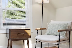 Ein Zimmer mit einem Nachttisch aus Holz, einem Stuhl mit Kissen und einer Stehlampe.