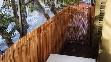 Un pequeño balcón con una mesa blanca, rodeado por una valla de madera, con vistas a una calle con árboles y edificios.
