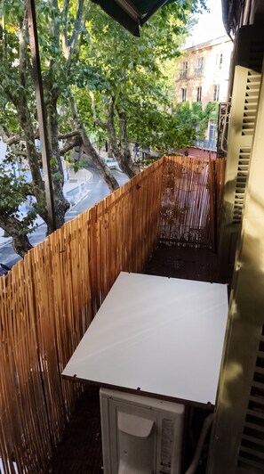 Un petit balcon avec une table blanche, entouré d’une clôture en bois, donnant sur une rue bordée d’arbres et d’immeubles.