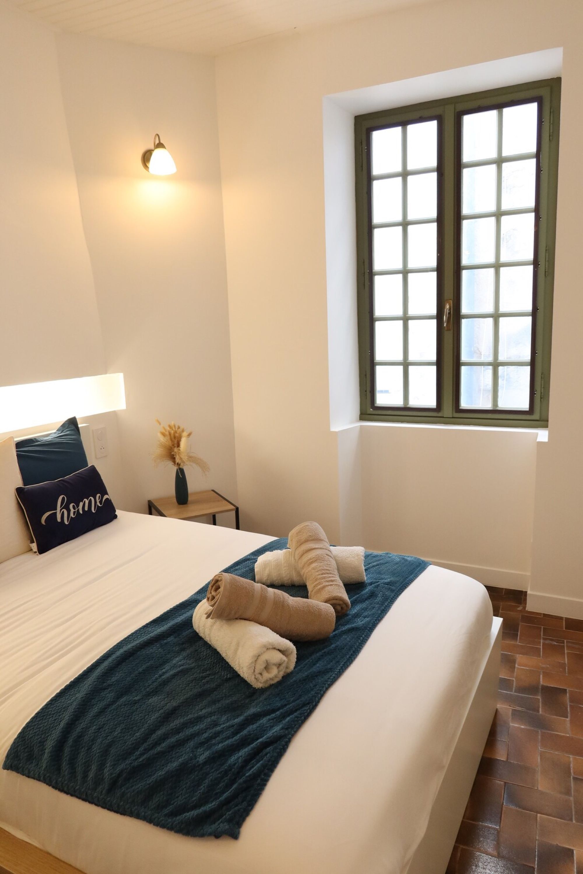 A neatly made bed with a blue blanket and rolled towels, a bedside lamp, a pillow with the word 'home,' and a window with a view.