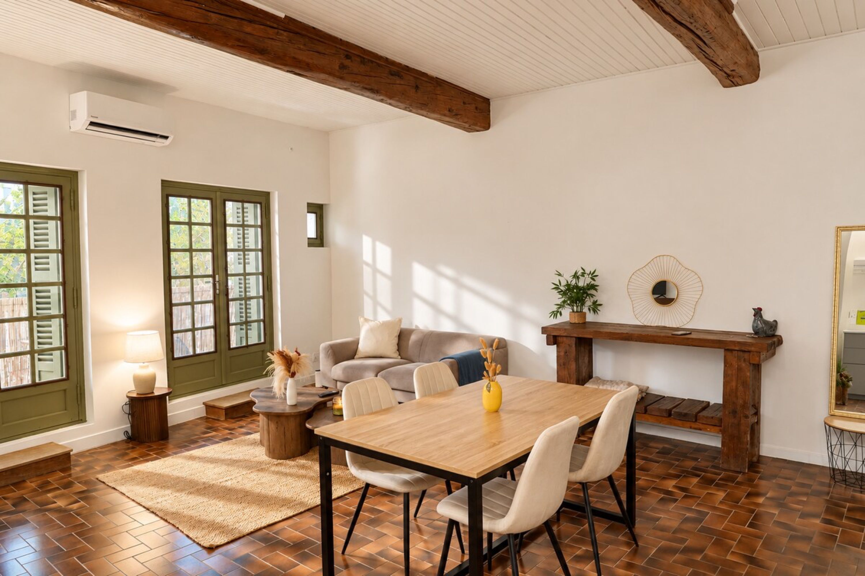 Un coin repas avec une table et des chaises en bois, une petite table d’appoint avec une lampe, et une vue sur l’extérieur par une porte à volets.