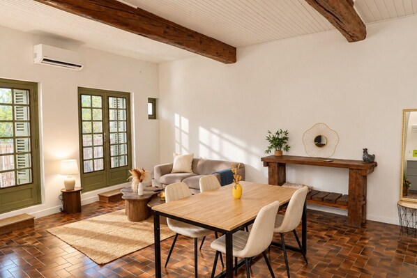 Un coin repas avec une table et des chaises en bois, une petite table d’appoint avec une lampe, et une vue sur l’extérieur par une porte à volets.