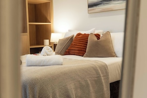 A hotel room with a bed, a bedside table with a lamp, and a wooden shelf. - West Kirby Apartment Walk to Marine Lake & beach (West Kirby)