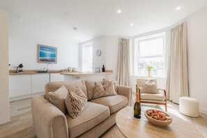 Ein modernes Wohnzimmer mit einem beigefarbenen Sofa, einem Esstisch mit einer Schüssel Essen, einer Weinflasche und einem Stuhl.