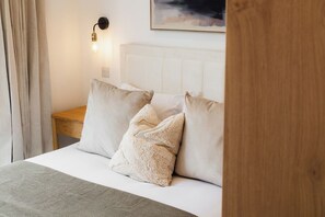 Ein Bett mit weißer Bettwäsche und zwei Kissen, ein Nachttisch aus Holz, eine Wandleuchte und ein Gemälde über dem Bett.