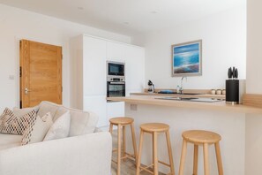 Une cuisine moderne avec une porte en bois, des placards blancs, un four à micro-ondes et un évier. On y trouve trois tabourets en bois et un canapé blanc agrémenté de coussins à motifs.