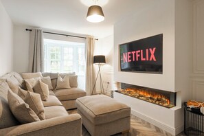 A living room with a beige sofa, ottoman, and a fireplace with a TV above it displaying 'NETFLIX'. - 3-Bed Chester Home | Parking | Near Chester Zoo (Cheshire West and Chester)